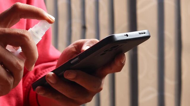 Asian man's hand spraying sanitizer and wiping a smartphone screen with a microfiber cloth. Cleaning and disinfecting a mobile phone to remove germs and bacteria.