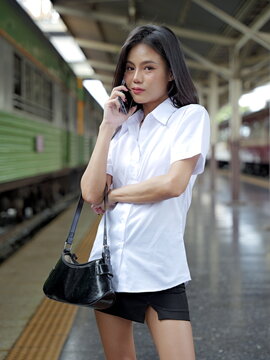 A young Asian woman wearing a Thai university student uniform stands on a train station platform talking on her smartphone