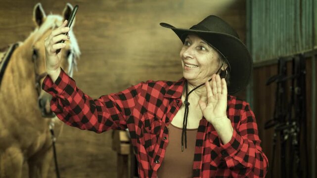 Senior cowgirl taking a selfie with smartphone inside a horse stable beside a palomino horse. Rural lifestyle, social media and western culture in 4K.