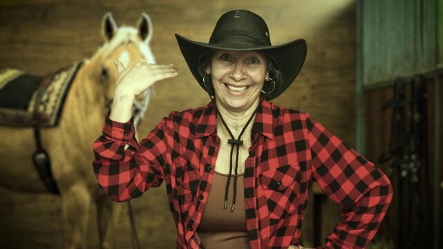 Smiling senior cowgirl making a playful hand gesture beside a saddled palomino horse inside a stable. Western lifestyle and rural authenticity in 4K.