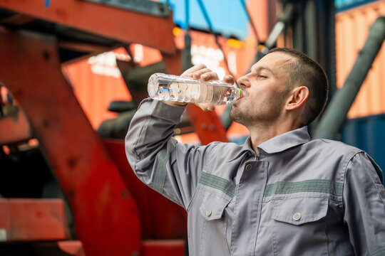 Thirsty industrial worker drinking fresh water from plastic bottle at container yard, Factory worker in workwear taking a break to drink water outdoors, Technician drinking mineral water from bottle