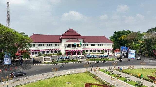 Smooth aerial orbit around Malang City Hall in East Java, Indonesia, featuring youth scout units practising with flags beside the historic Dutch colonial building.