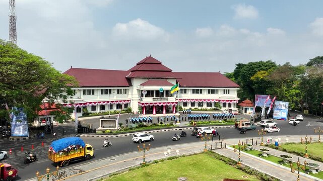 Aerial footage approaching historic Malang City Hall in East Java, Indonesia, as youth scout units practise with flags outside the Dutch colonial landmark on a sunny day.