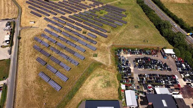 Rows of solar panels line open farmland beside sprawling vehicle junkyard and industrial warehouses, highway running past dense tree line in rural countryside nearby. Aerial drone top down flyover