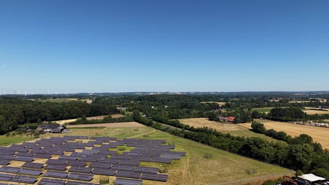 ast solar panel field stretches beneath wide open blue sky toward wooded countryside, distant wind turbines and red-roofed farmhouse visible near dense green tree line. Aerial drone rising wide shot