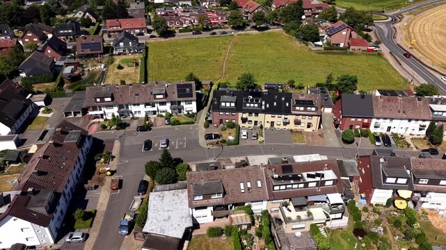 Rows of terraced houses with solar panel roofs line quiet residential streets in small German town, green fields and golden wheat crops surrounding neighborhood below. Aerial drone lateral wide shot