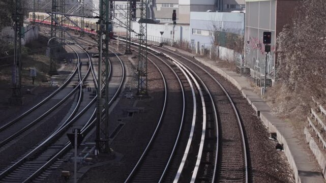 An elevated view shows a S-Bahn train traveling along railway tracks in an urban setting. Steel overhead power masts stand in the foreground as the train curves along the rails on a clear day.