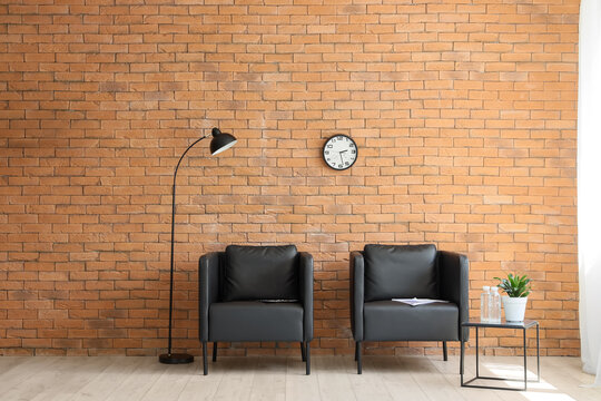 Empty waiting room with two black armchairs, a floor lamp, and a clock on a brick wall, modern office interior