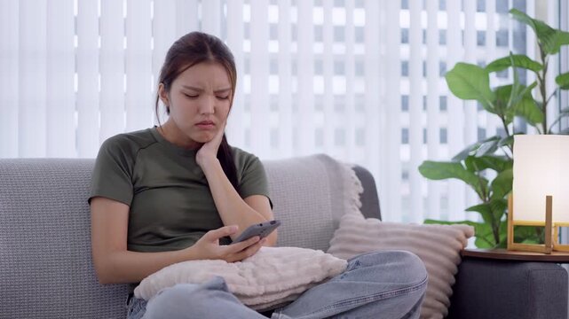 Young Asian woman feeling stressed and anxious while using smartphone on living room sofa
