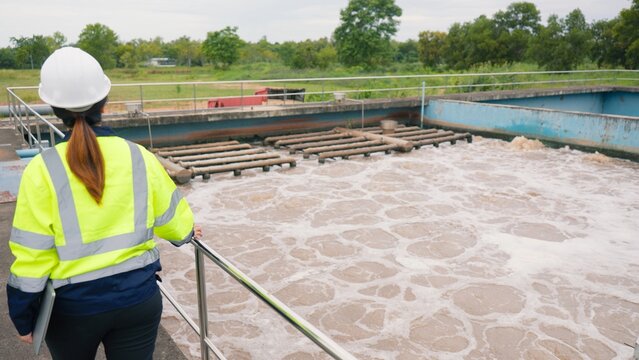 Asian female engineer technician in safety helmet inspecting wastewater treatment plant facility using laptop computer. Environmental engineering and water management concept.