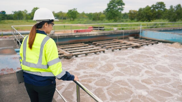 Asian woman worker checking industrial water treatment facility and wastewater basin with laptop and clipboard. Environmental protection, sanitation and engineering concept.