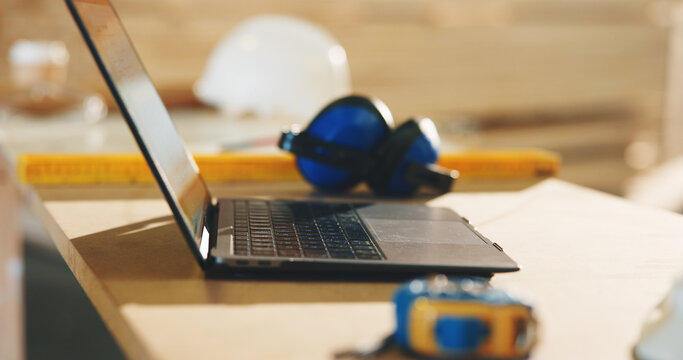 Carpentry, laptop and earmuffs on desk in workshop for hearing protection or safety equipment. Woodwork, technology and computer for order of timber project or furniture manufacturing in factory.