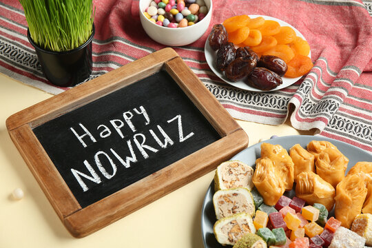 Chalkboard with text HAPPY NOWRUZ, traditional sweets and green wheat grass on yellow background, closeup. Nowruz Bayram celebration