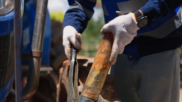 Asian technician worker using grease gun tool lubricating heavy machinery tractor joint parts at farm site. Professional industrial mechanic service and vehicle maintenance inspection.