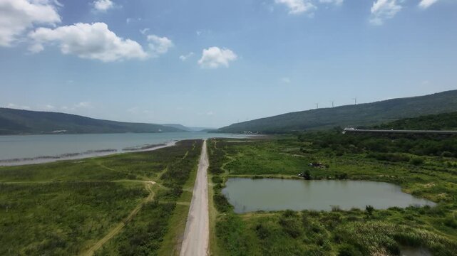 Empty dirt road leads to a vast, blue lake surrounded by mountains. Pak Chong, Thailand.