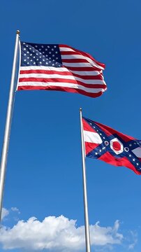 American and Ohio Flags Waving Against Blue Sky - Two flags, the American flag and the Ohio state flag, are depicted fluttering in the wind against a clear blue sky.