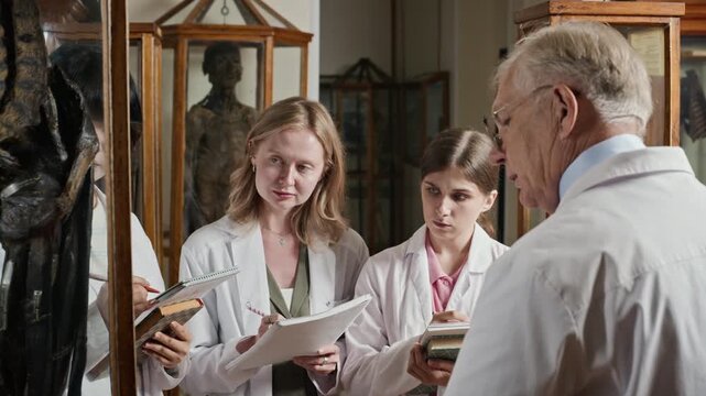Senior Caucasian male professor explaining anatomical specimen to diverse young adult female medical students in museum. Perfect for medical school promotions or healthcare training videos