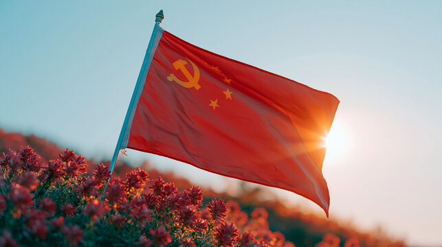 Chinese flag waving in wind against blue sky and red flowers illuminated by sun.