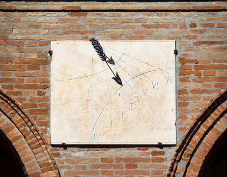 Ancient sundial with metal gnomon shaped like arrow mounted on brick wall in Mondaino Italy showing Roman numerals and sunlight shadow lines