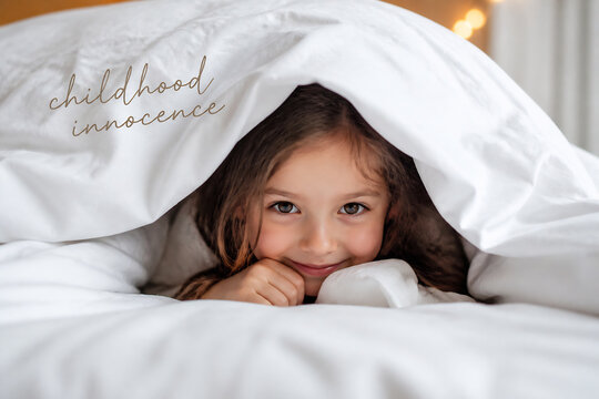 A young girl peeking out from under white bedding in a bright room.