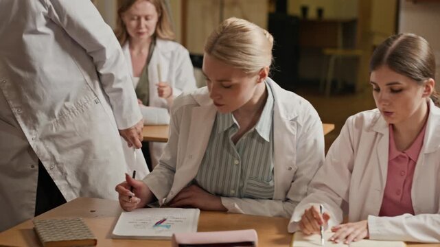 Diverse young adult female medical students in lab coats writing in notebooks during lecture, camera panning. Video is ideal for medical school promotions and healthcare education marketing