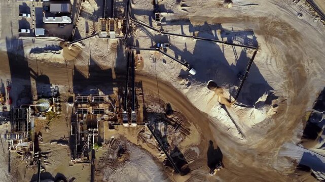 Drone video captures a straight-down, sliding aerial of an active gravel and sand pit at sunrise. Long shadows emphasize machinery, conveyors, and earth textures in crisp golden light.