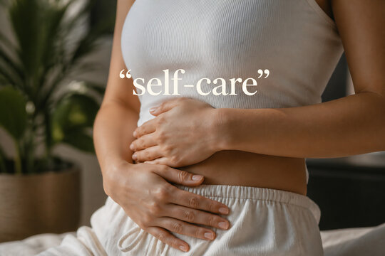 A person in a white top and pants is holding their stomach indoors, suggesting self-care.