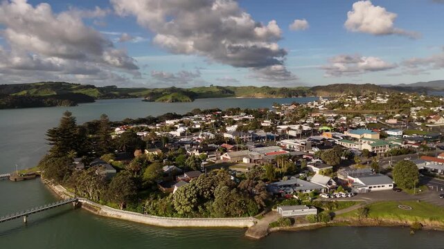Peninsula town clusters along waterfront pier near helicopter pad at Raglan, New Zealand, distant islands and green hills stretch across calm bay beneath cloudy blue sky. Aerial drone orbit wide shot