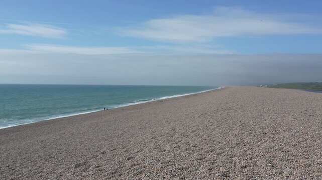 Sweeping panoramic shot of Chesil Beach near Weymouth, featuring endless pebbles, calm blue sea, and clear sky in bright daylight. No people visible.