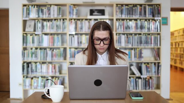 University student works on her thesis in a library using a laptop researching academic sources and writing her final graduation project with deep focus before submission