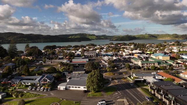 Cars circle busy roundabout intersection near town shops and helicopter pad at Raglan, New Zealand, hillside houses and estuary water stretch beneath partly cloudy sky. Aerial drone wide shot