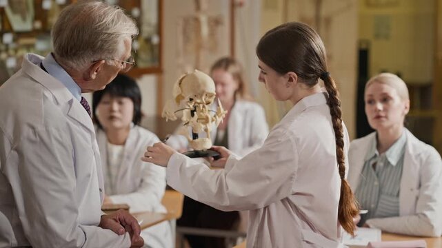 Young adult Caucasian female medical student pointing at human skull model during anatomy class with senior Caucasian professor, zoom out. Ideal for medical school marketing