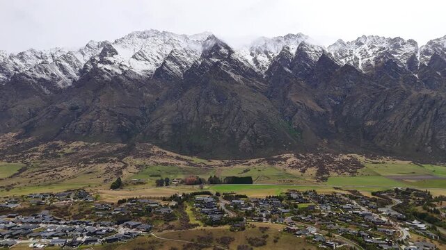 Aerial drone footage sliding left across Jack&rsquo;s Point, revealing residential neighborhoods, open grasslands, and dramatic snow-capped alpine mountains in Queenstown, New Zealand.