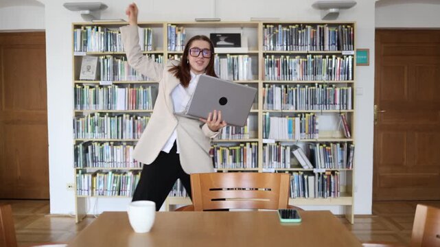 Excited office employee celebrates a successful business deal while looking at her laptop and dancing with joy after receiving positive results from her work