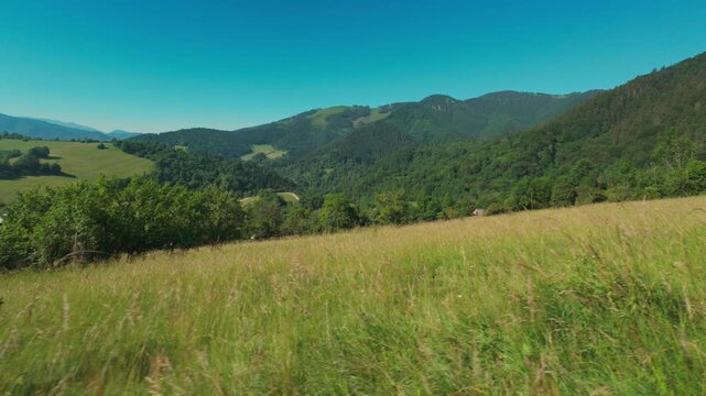 POV sliding shot moving left to right along a mountain path with views of green meadows and forested hills under a clear blue sky