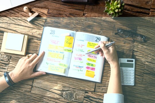 Person writing in a daily planner with colorful sticky notes on a wooden desk calendar schedule organizer