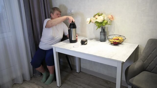 Senior woman sitting at table in domestic setting, opening thermos, preparing drink. Kitchen area with white table, basket of fruit, fresh flowers in vase, window curtain, comfortable chair.