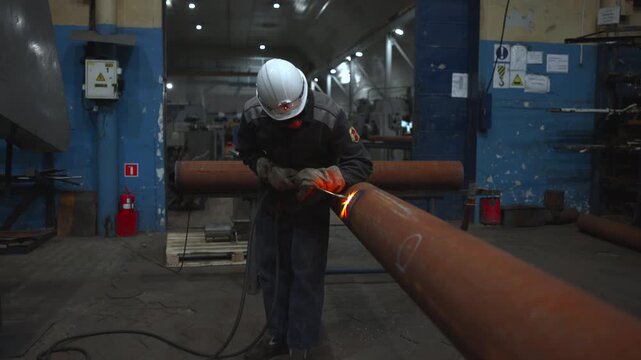 Industrial worker cutting rusted steel pipe with handheld torch in workshop, sending bright orange sparks and molten droplets across worn metal surface under dim overhead lighting