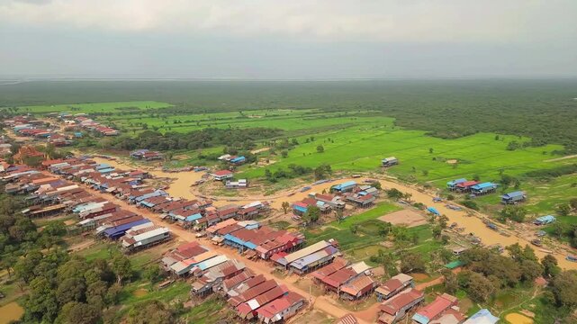 Cinematic 4K aerial drone footage of Tonle Sap floating village in Cambodia, showcasing a winding muddy canal lined with traditional stilt houses, colorful fishing boats, lush green countryside