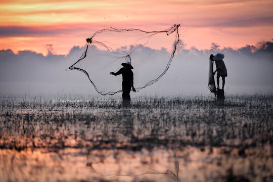 Silhouette of traditional fishermen casting net at golden sunrise in Sakon Nakhon, Isan, Thailand. Capturing authentic rural Asian lifestyle, culture, and fishing for daily sustenance in tranquil lake