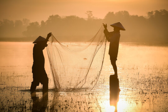 Silhouette of traditional fishermen casting net at golden sunrise in Sakon Nakhon, Isan, Thailand. Capturing authentic rural Asian lifestyle, culture, and fishing for daily sustenance in tranquil lake