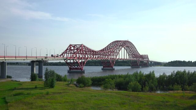 Red arched steel truss bridge spanning wide river with tanker truck on approach road under pale blue sky and calm daylight, showing strong industrial structure, open landscape, and long cross-river