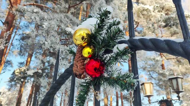 Christmas decoration detail on a ornate metal fence, showcasing snow-covered fir branches, pine cone, and vibrant red and gold baubles against a backdrop of frosted pine trees and clear winter sky.
