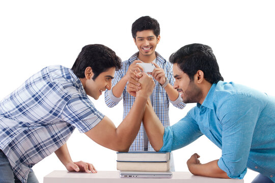 Two friends arm wrestling while other friend taking photo