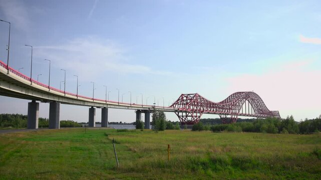 Elevated concrete highway curving into red steel truss arch bridge over river with grassy meadow foreground and pale blue sky in panoramic scenic landscape, with bridge, truss, highway, arch detail