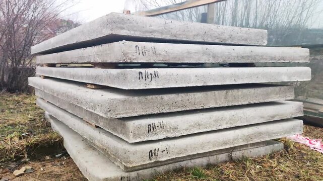 Industrial supply of heavy gray concrete slabs for construction purposes, neatly stacked on ground outdoors with dry grass and bare trees in background under cloudy sky, material