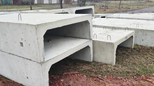 Precast concrete storm drain sections stacked outdoors on construction site ground, featuring rectangular shapes, rebar hooks for lifting, rough texture of material, and overcast sky