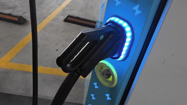 Electric vehicle charging plug connected to public charging station with illuminated blue light ring in parking garage, representing future of sustainable transportation and green energy.