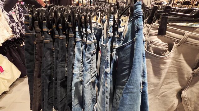 Variety of denim trousers and pants in blue and grey hues displayed on clothing rack in retail store interior, presenting diverse textile textures and shades for consumer selection
