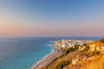 Panoramic high-angle view of Psaropoula Beach and Rhodes Town at sunset, looking towards the northern tip of Rhodes Island, Greece. Turquoise Aegean Sea, coastline, and city architecture bathed in gol © Artem